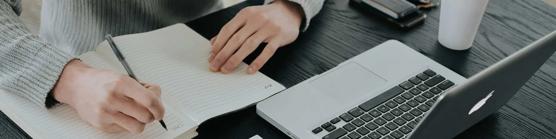 Dokumentation: Hand schreibt Notizen in ein Notizbuch, daneben ein aufgeklappter Laptop