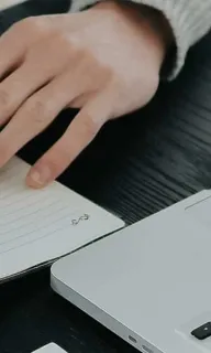 Dokumentation: Hand schreibt Notizen in ein Notizbuch, daneben ein aufgeklappter Laptop