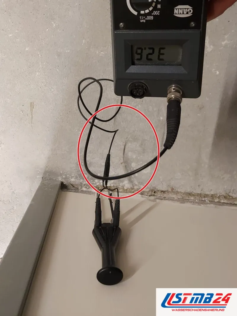 Messgerät mit Kabel zu zwei Elektroden am Rand eines Estrichs neben der Wandabdichtung, die Hand am Gerät, ein roter Kreis markiert die Stelle