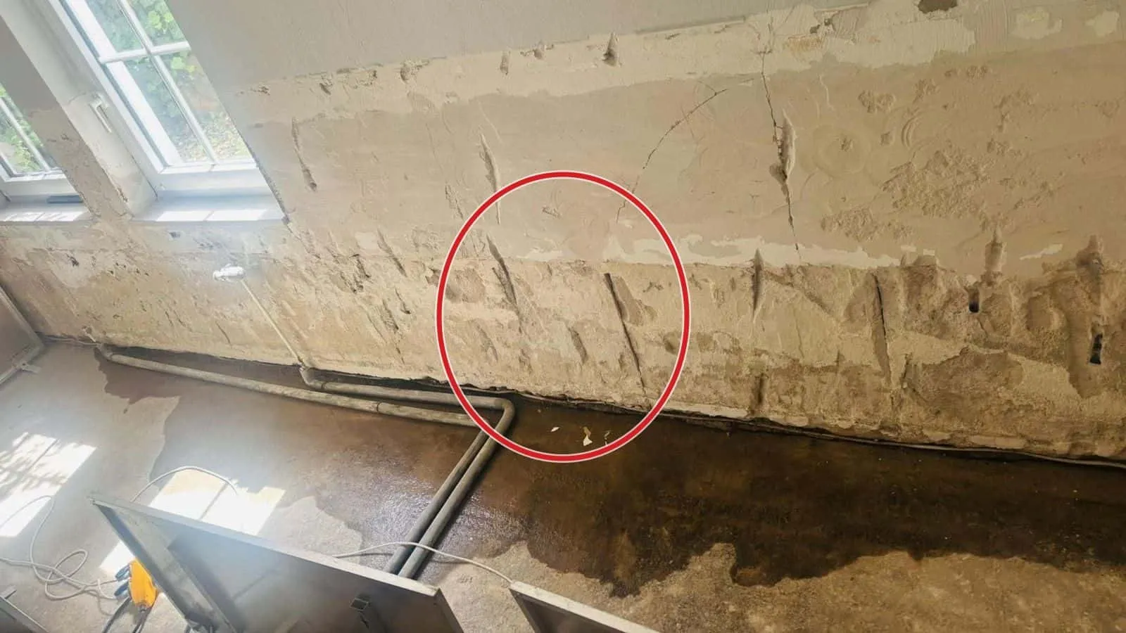 Kellerwand mit abgeschlagenem Putz, nasser Boden und Heizungsrohre unter dem Fenster