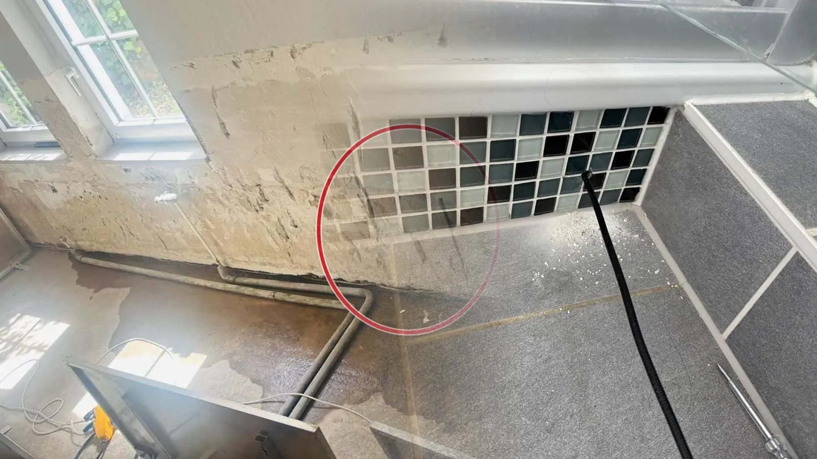 Bad mit Glasmosaik an der Wanne, ein schwarzer Trocknungsschlauch steckt in einer Bohrung in der Bodenfuge