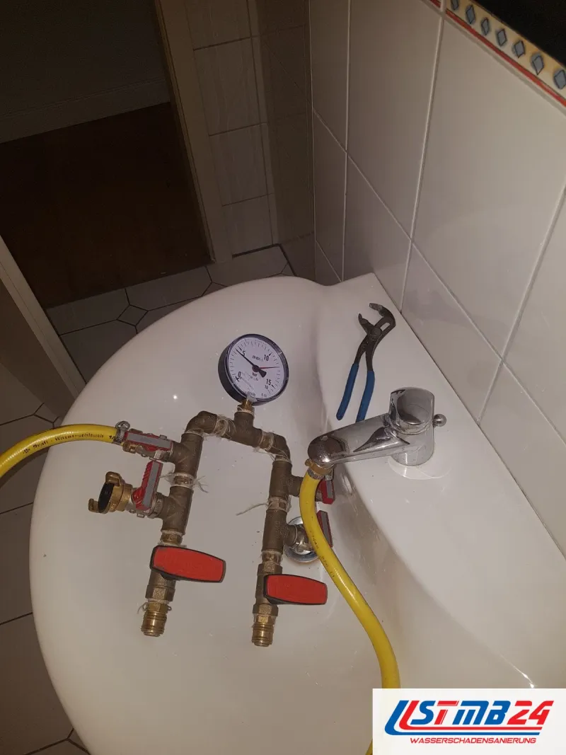 Druckprüfgerät mit Manometer und roten Hähnen auf einem Waschbecken, daneben eine Rohrzange und ein gelber Schlauch