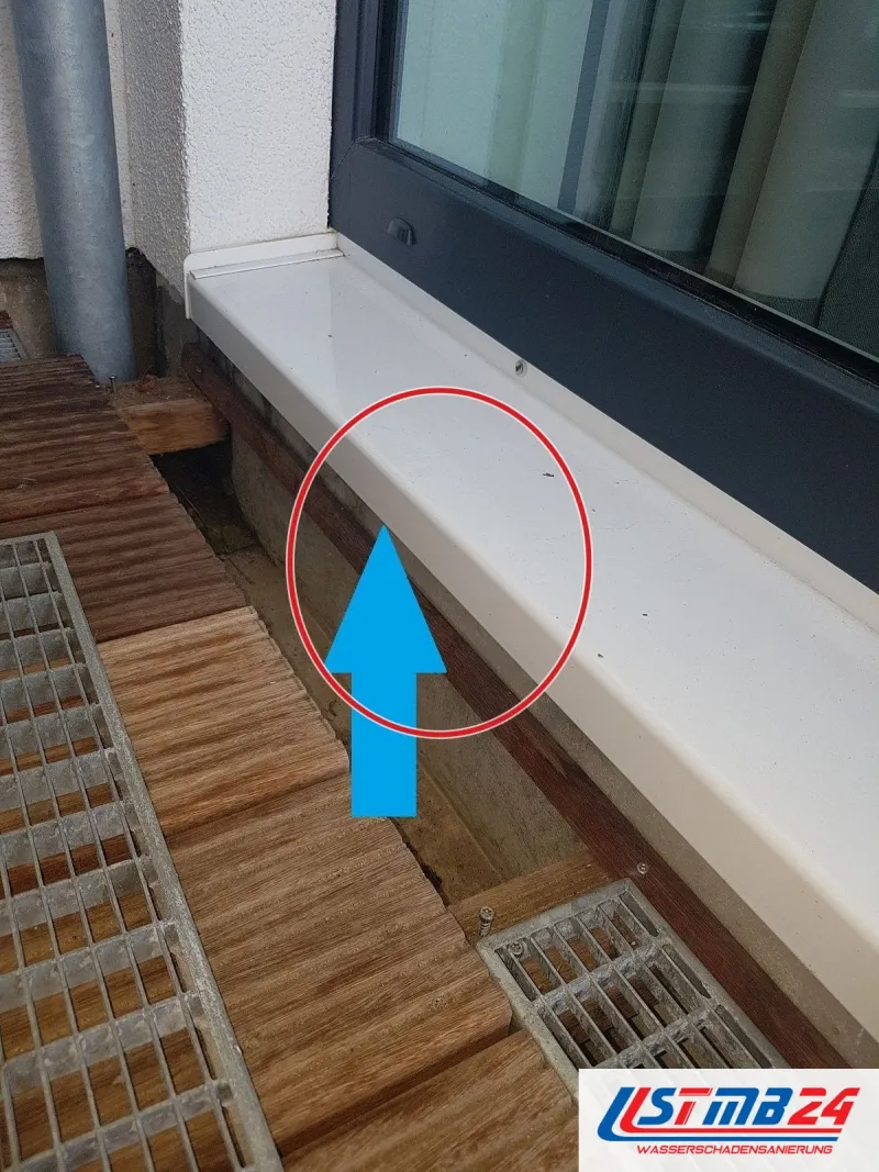 Weiße Fensterbank außen mit blauem Pfeil auf die offene Fuge zur Fassade, Holzterrasse und Gitterrost darunter, ein roter Kreis markiert die Stelle