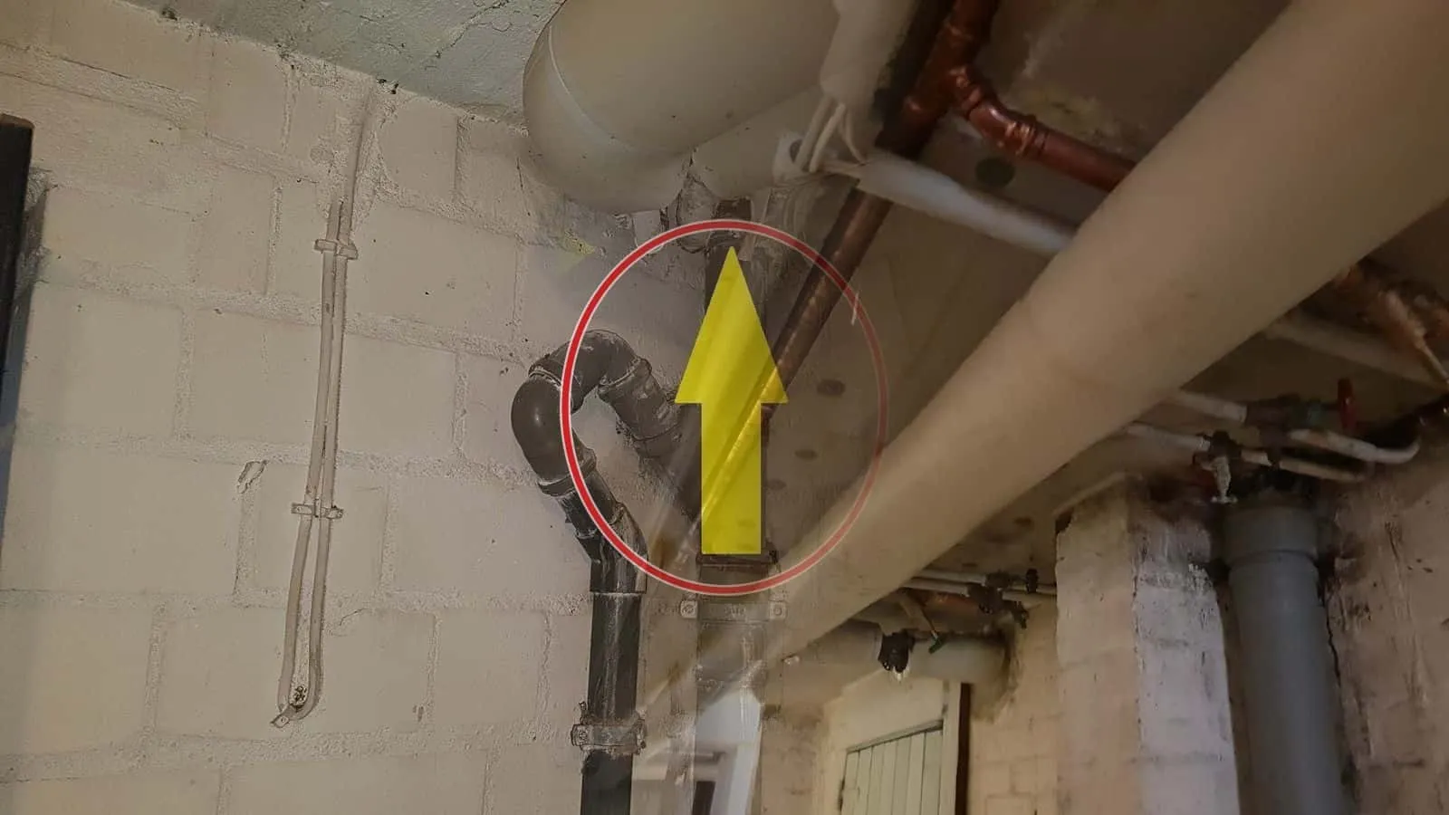 Kupfer- und Abflussleitungen unter einer Kellerdecke, ein gelber Pfeil zeigt auf einen Fleck
