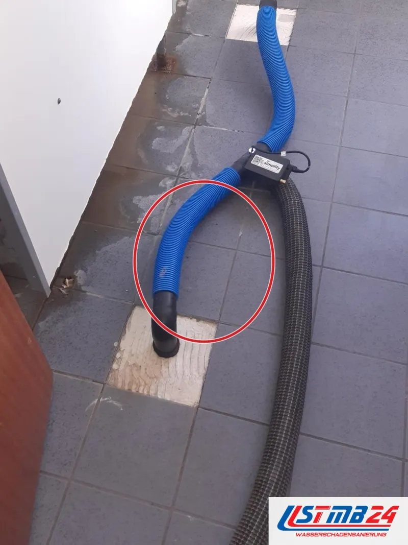 im Flur ist eine Fliese herausgenommen, der schwarze Saugstutzen sitzt in der Bohrung, ein roter Kreis markiert die Stelle