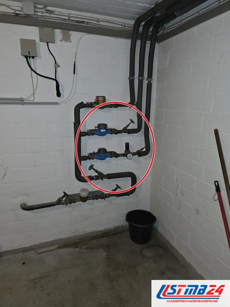 Hausanschluss mit drei Wasserzählern übereinander, gedämmte Leitungen und Absperrventile an weißer Kellerwand, ein roter Kreis markiert die Stelle