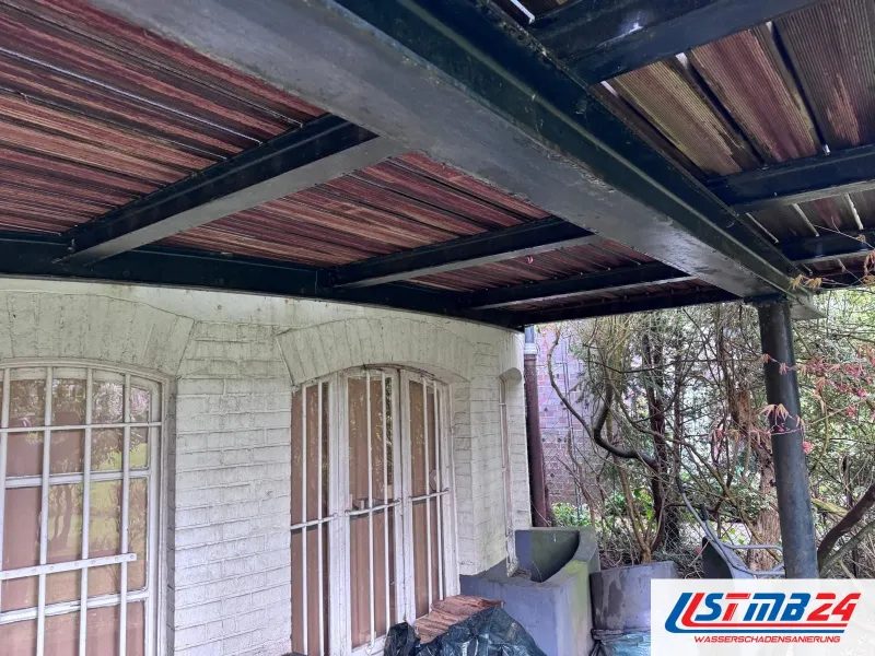 Überdachte Terrasse vor einer weiß geschlämmten Klinkerwand mit vergitterten Rundbogenfenstern, darüber Stahlträger und Platten, daneben Sträucher