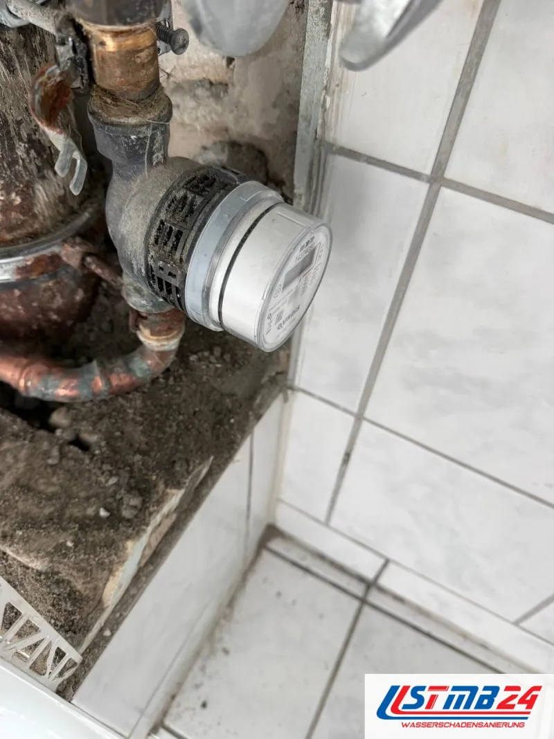 Wasserzähler an einer Leitung in einer offenen Bodenecke neben weißen Fliesen, Verschraubungen mit Korrosion, darunter Erde und ein Kupferrohr