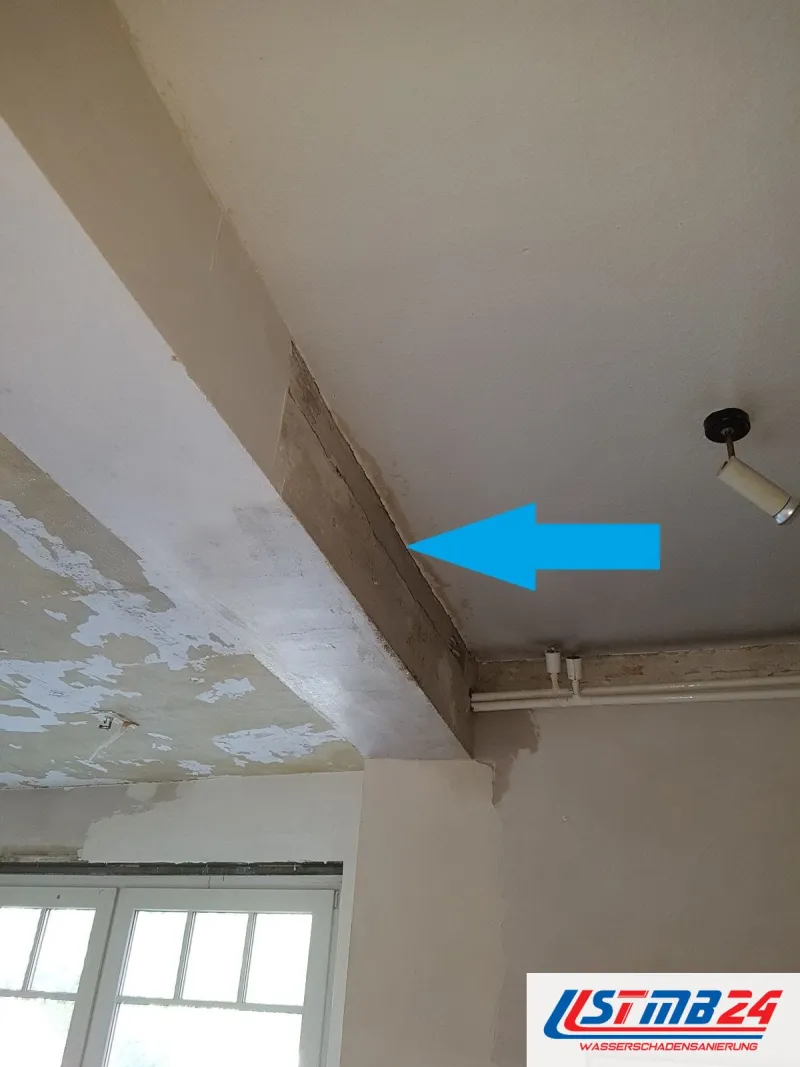 Zimmerdecke mit einem Unterzug, ein blauer Pfeil zeigt auf die feuchte Fuge zwischen Unterzug und Decke, unten ein Sprossenfenster