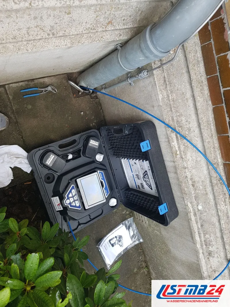 Geöffneter Koffer mit Monitor im Hof neben einem grauen Fallrohr, das blaue Schubkabel führt in den Anschluss, vorn Sträucher