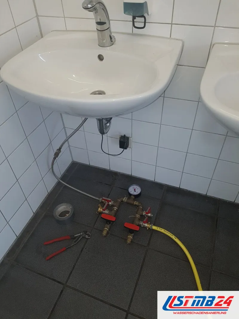 Waschbecken an einer weiß gefliesten Wand, am Eckventil ein Prüfgerät mit Manometer, auf dem dunklen Boden Werkzeug