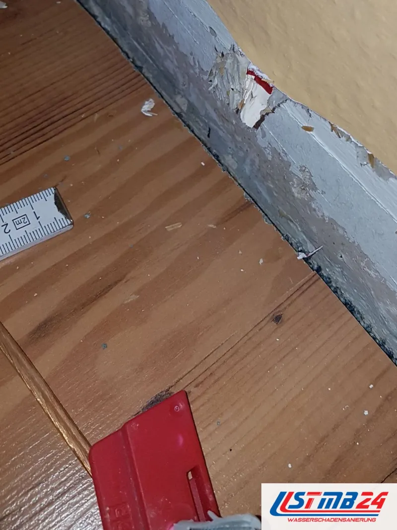 Dielenboden an einer Wand ohne Sockelleiste, der Putz ist aufgeweicht, ein Zollstock liegt als Maßstab daneben