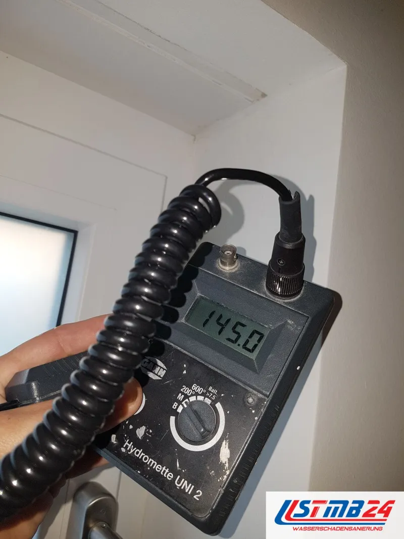 Feuchtemessgerät mit 145,0 auf dem Display an der Wand neben einem Türrahmen, das Spiralkabel hängt herab