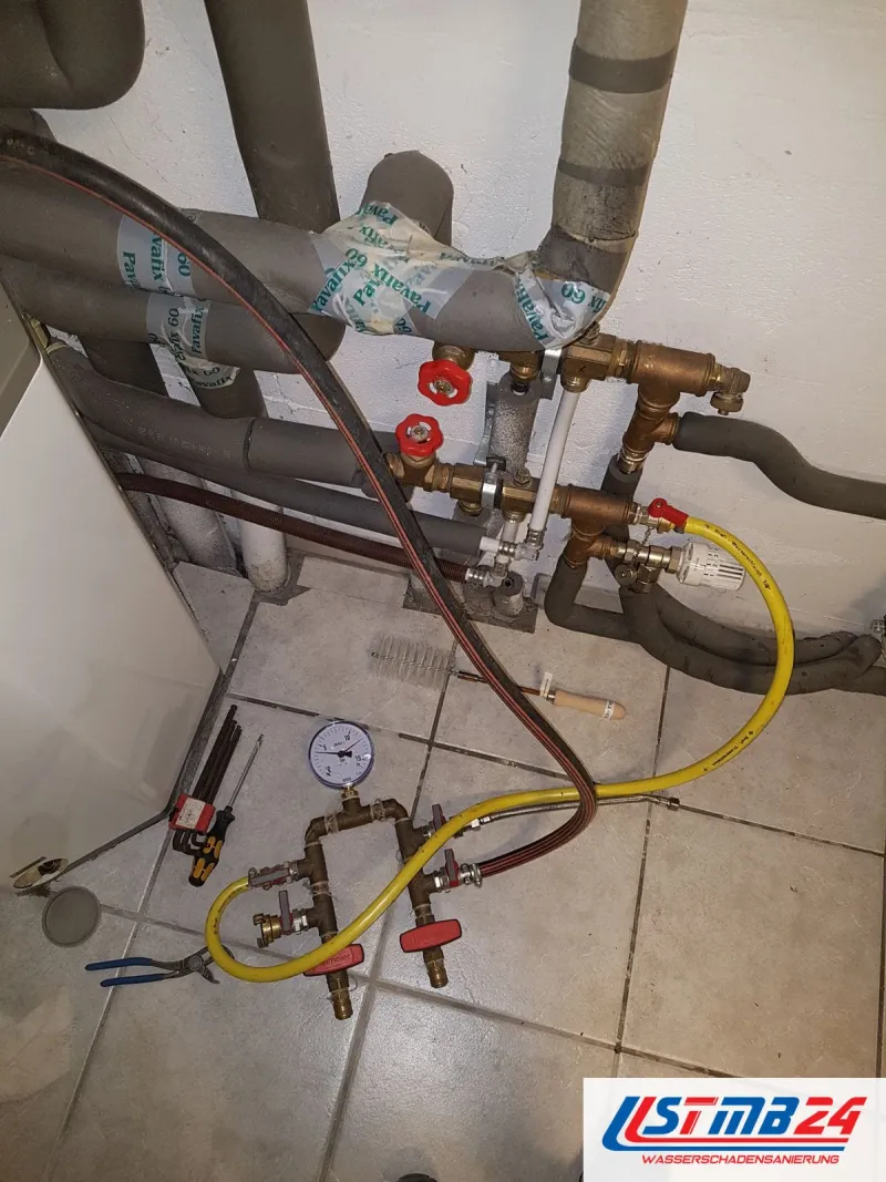 Prüfgerät mit Manometer und gelbem Schlauch an den Heizungsleitungen im Hausanschlussraum, auf dem Fliesenboden Werkzeug