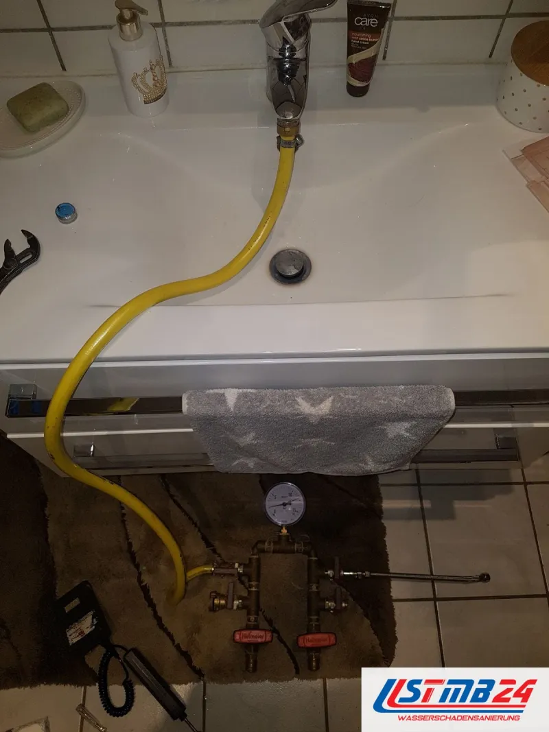 Waschtisch, vom Hahn führt ein gelber Schlauch hinunter zum Prüfgerät mit Manometer auf einem braunen Vorleger