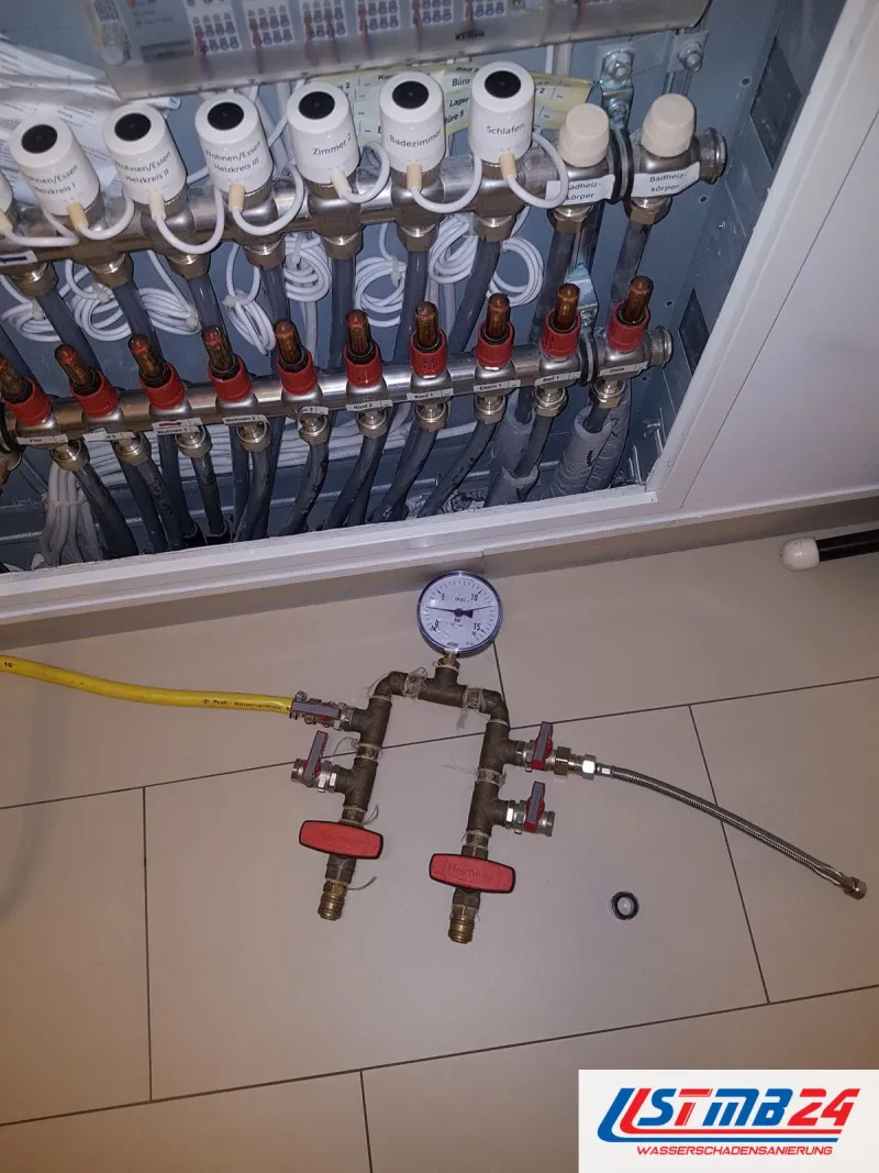Verteiler der Fußbodenheizung mit Stellventilen, davor ein Prüfgerät mit Manometer und roten Hähnen auf den Fliesen