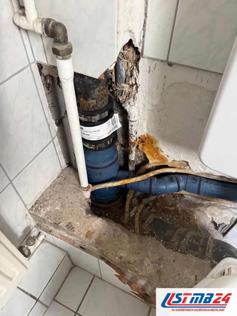 Aufgestemmte Ecke neben dem WC: altes Gussrohr mit neuem Übergang auf ein blaues Abflussrohr, daneben rostige Wasserleitungen