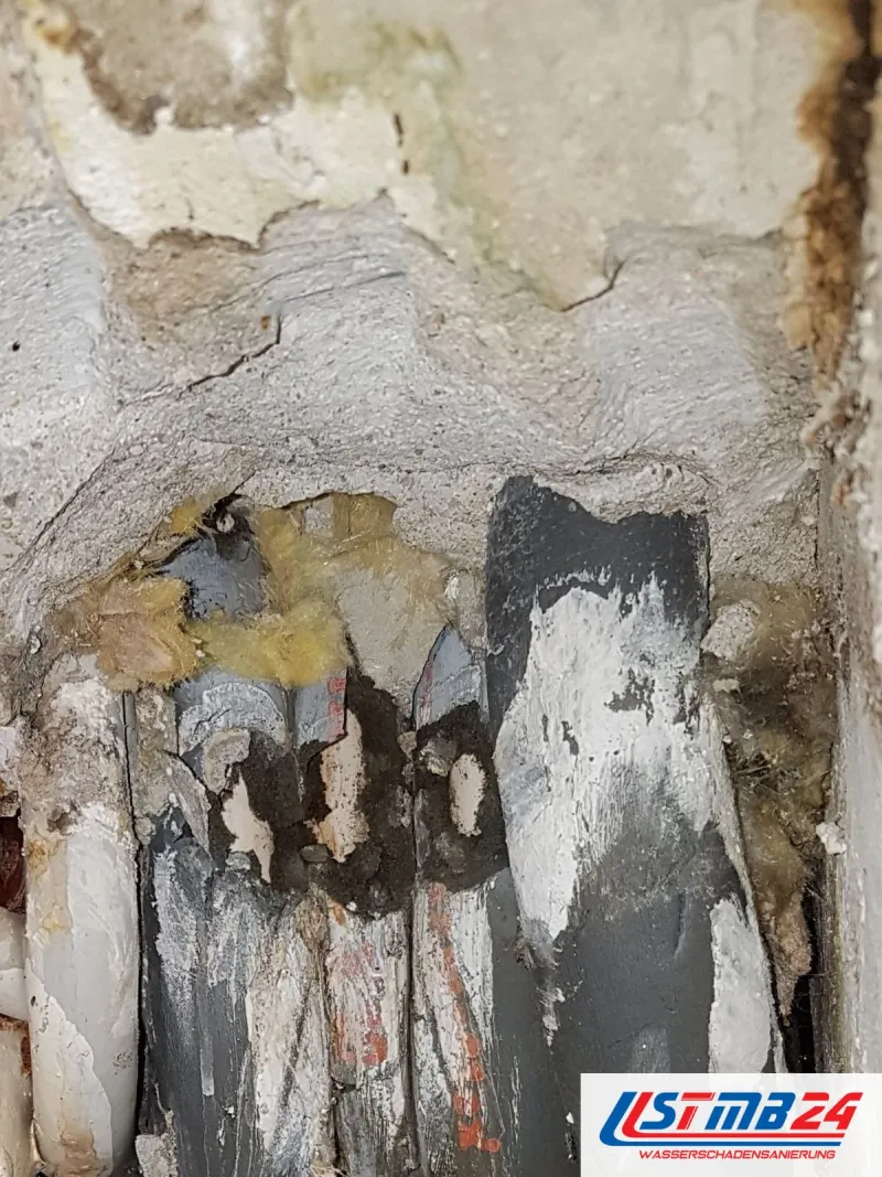 Ausschnitt einer Wand mit abgeplatztem Putz, darunter dunkle, rostige Rohre und Reste gelber Dämmung