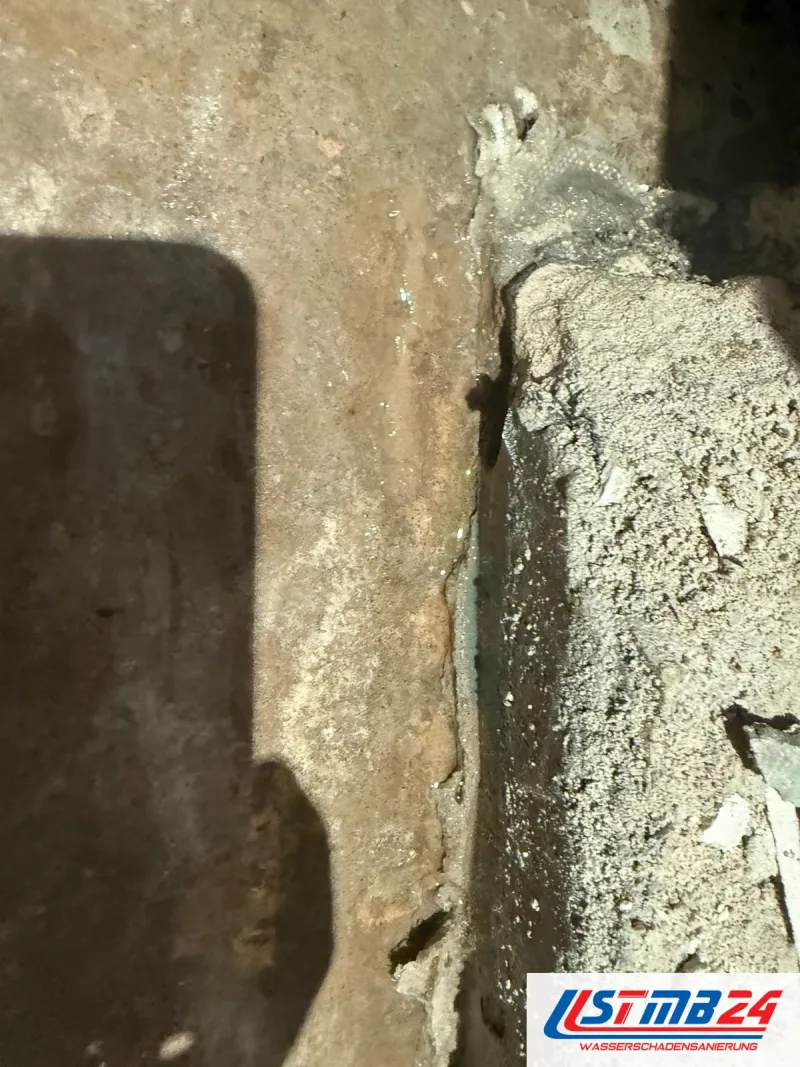 Aufgestemmter Estrich mit nasser Rinne entlang der Leitung, Wasser glänzt im Bruch, daneben loser Schutt