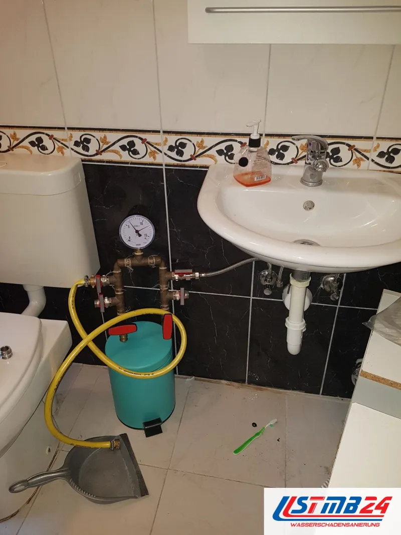Bad mit dunklen Wandfliesen und Bordüre, unter dem Waschbecken ein Prüfgerät mit gelbem Schlauch und ein grüner Kanister