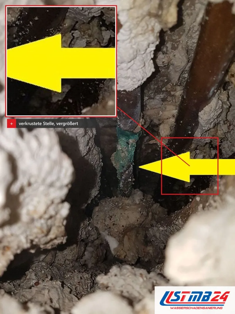 Enger Hohlraum in einer Wand mit alten Rohren, vergrößert eine grün verkrustete Stelle, auf die ein gelber Pfeil zeigt