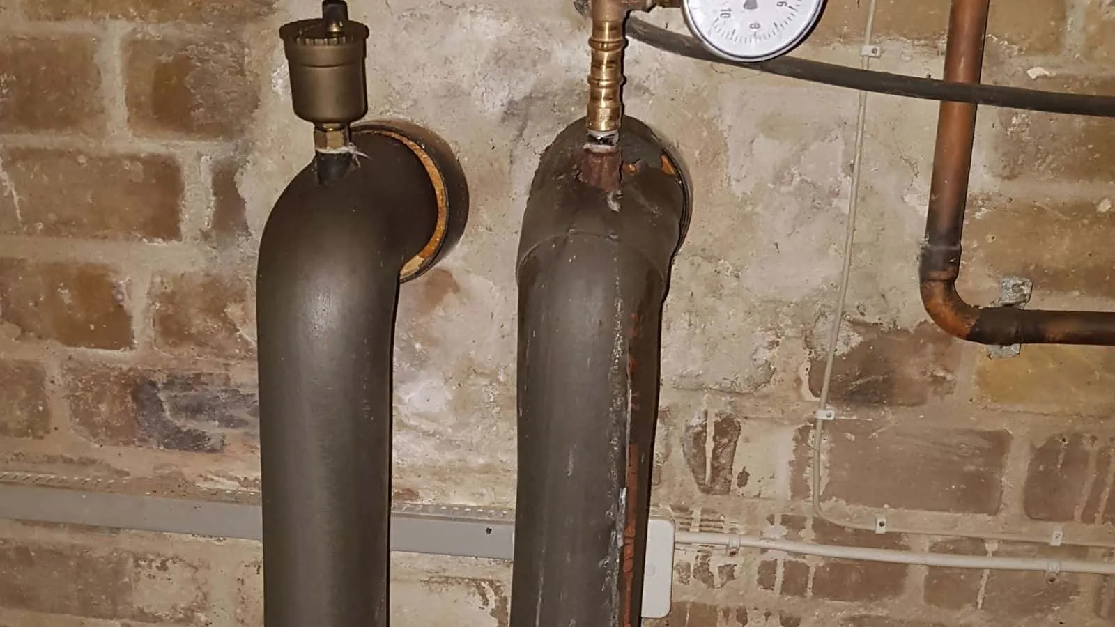 Zwei gedämmte Rohrbögen mit Entlüftern vor einer alten Klinkerwand, oben ein Manometer