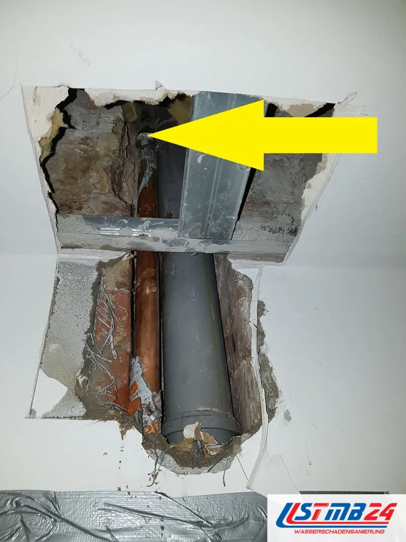 Geöffnete Decke mit Kupferleitung und grauem Fallrohr, ein gelber Pfeil zeigt auf die nasse Stelle, darunter Folie
