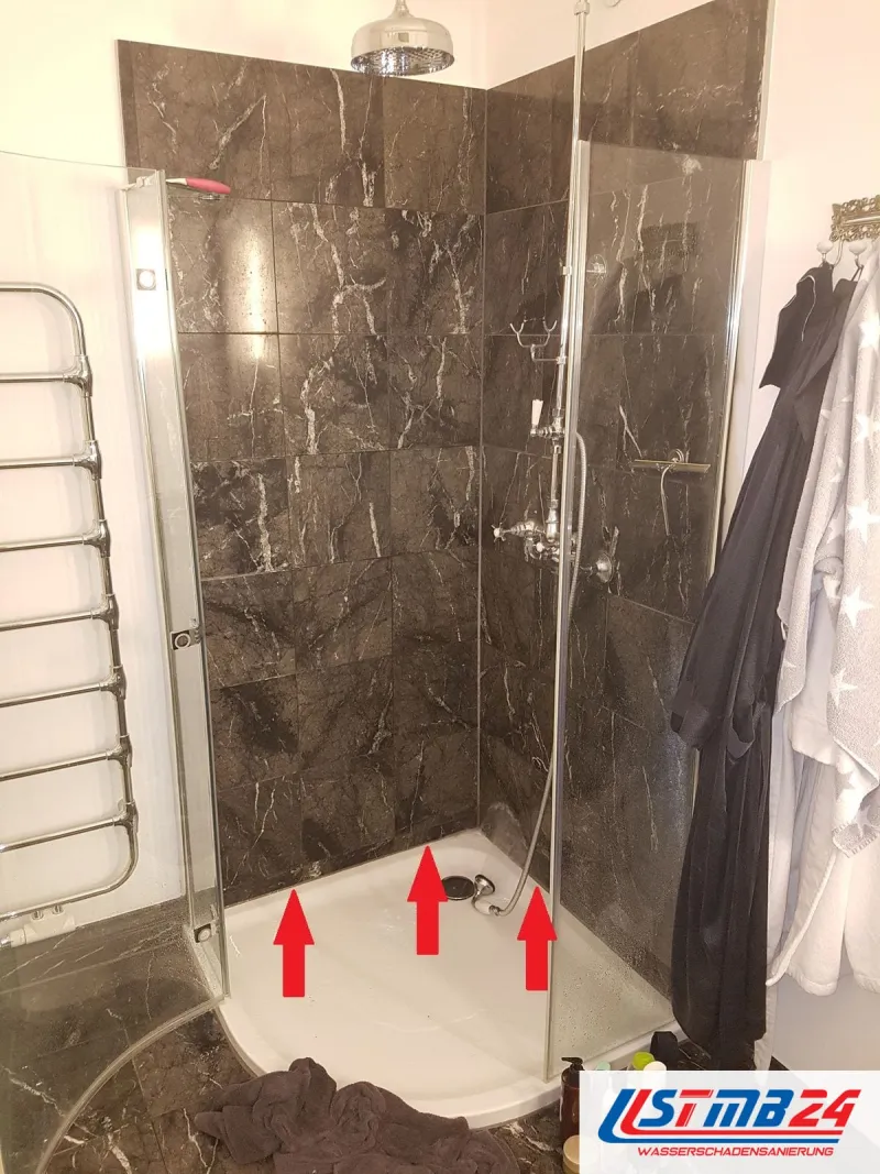 Dusche mit dunklen Marmorfliesen und Glastür, drei rote Pfeile zeigen auf die Fuge am Rand der Duschtasse