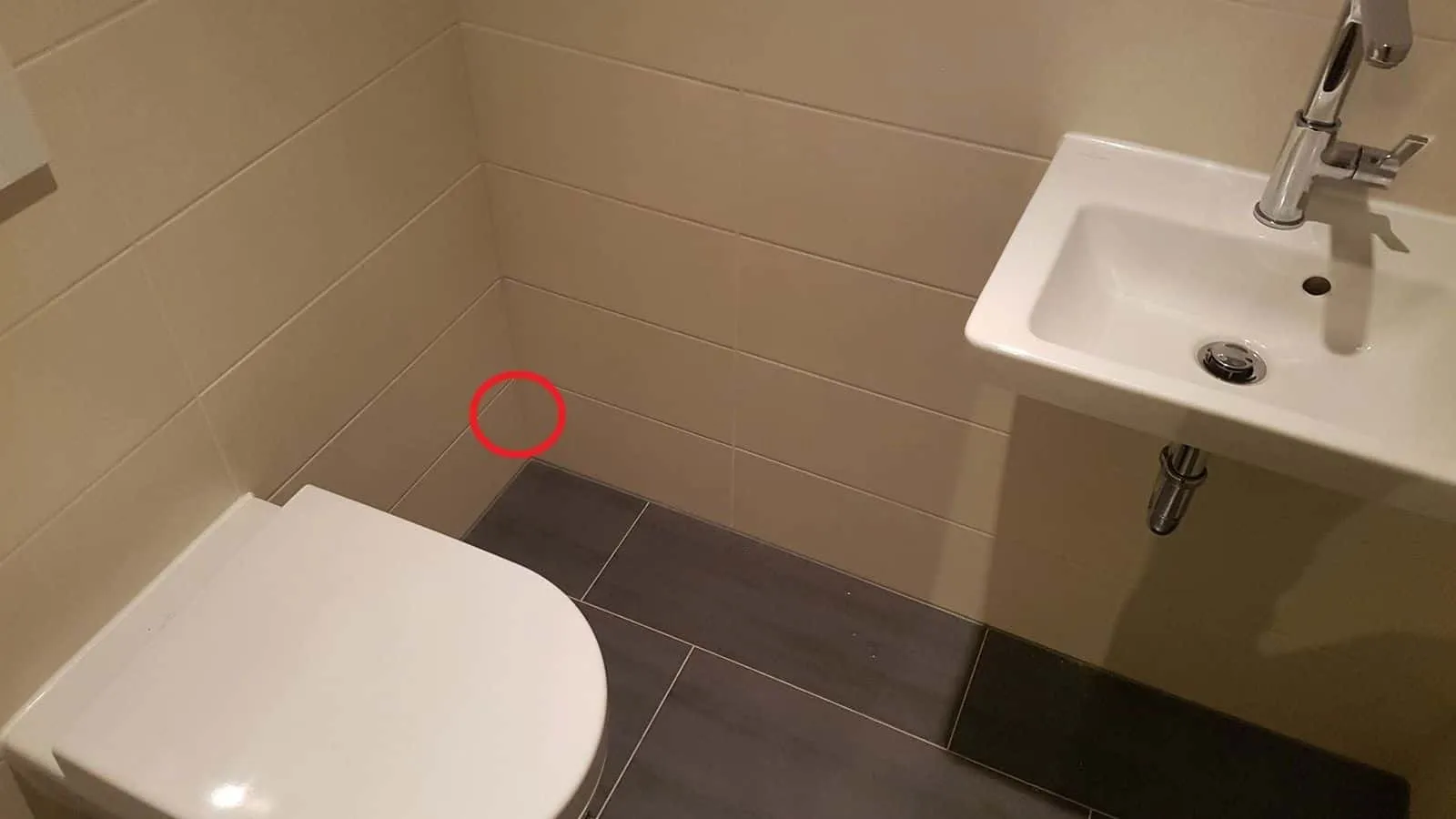 Kleines Bad mit wandhängendem WC und weißem Waschtisch, ein roter Kreis markiert eine Stelle an der Fliesenwand über dem WC