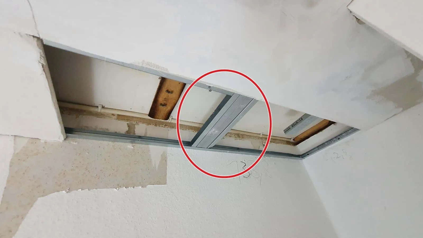Geöffnete Gipskartondecke, darüber Metallprofile, Holzbalken und eine Leitung, ein roter Kreis markiert die Stelle