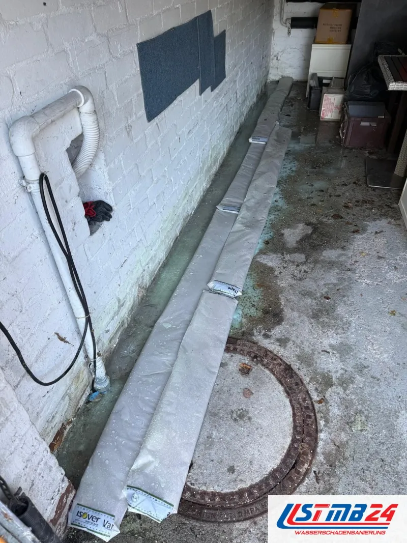 Kellergang mit nassem Boden und Schachtdeckel, an der Wand liegen zwei gefüllte Schlauchbarrieren gegen das Wasser