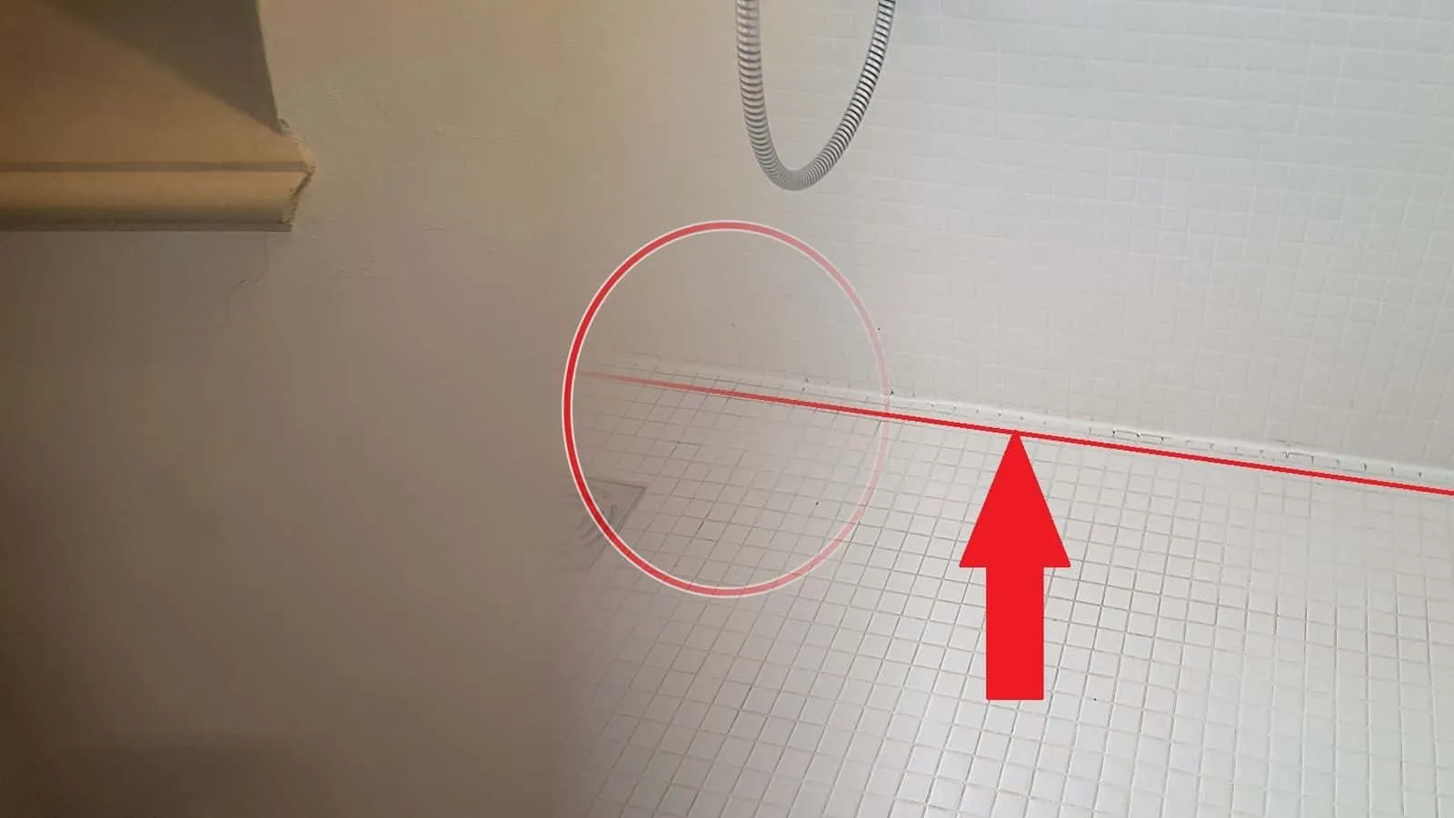 Weiße Mosaikdusche mit Bodenablauf, an der Fuge zwischen Wand und Boden eine rote Linie mit Pfeil