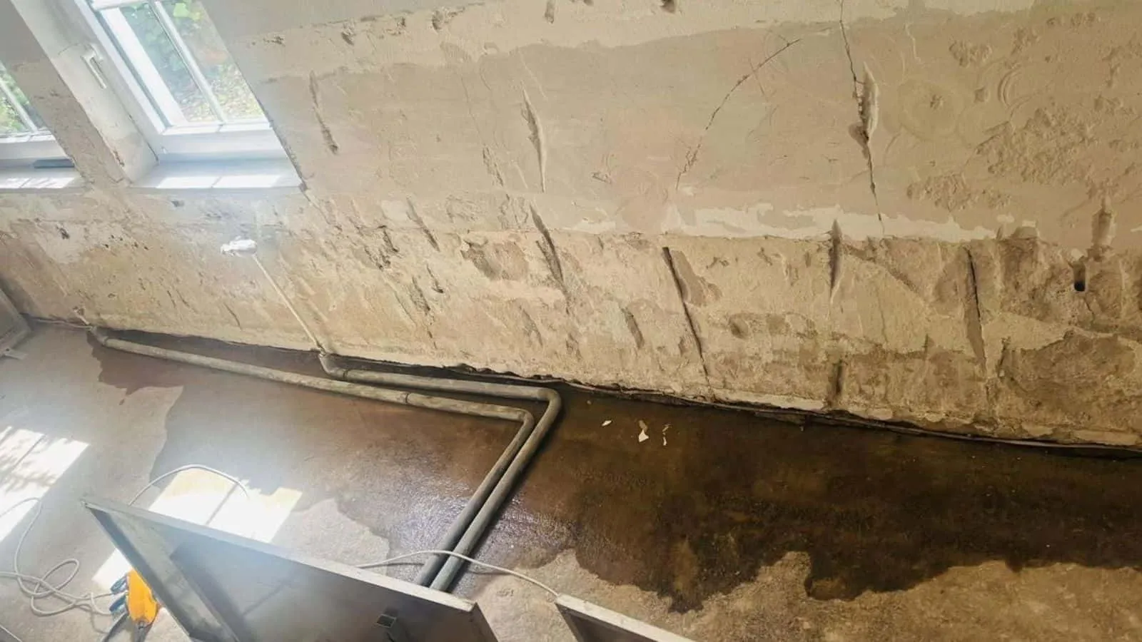 Kellerwand mit abgeschlagenem Putz, nassem Boden und Heizungsrohren am Fenster