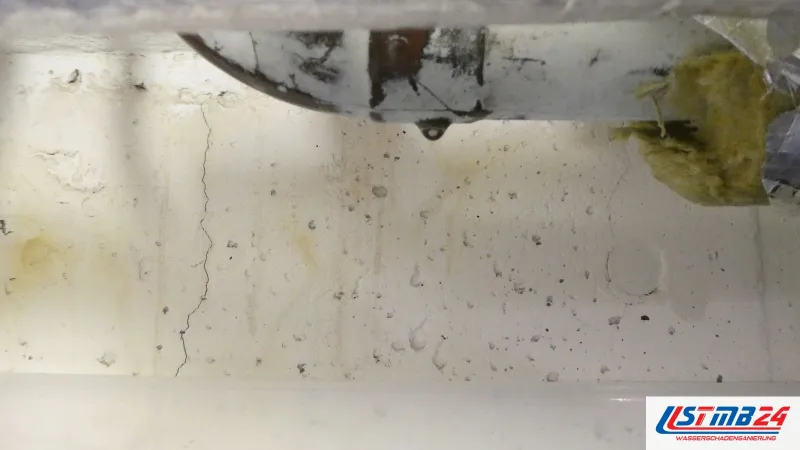 Rohre unter einer Kellerdecke, an der Leitung hängt ein Wassertropfen, darunter gelbe Laufspuren und ein Riss in der Wand