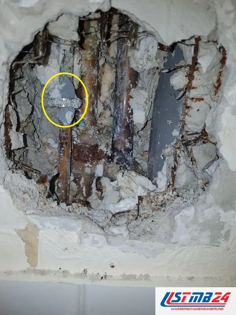 Aufgestemmte Wand mit stark verrosteten Stahlrohren neben einem grauen Abflussrohr, gelb umkreist eine umwickelte Flickstelle