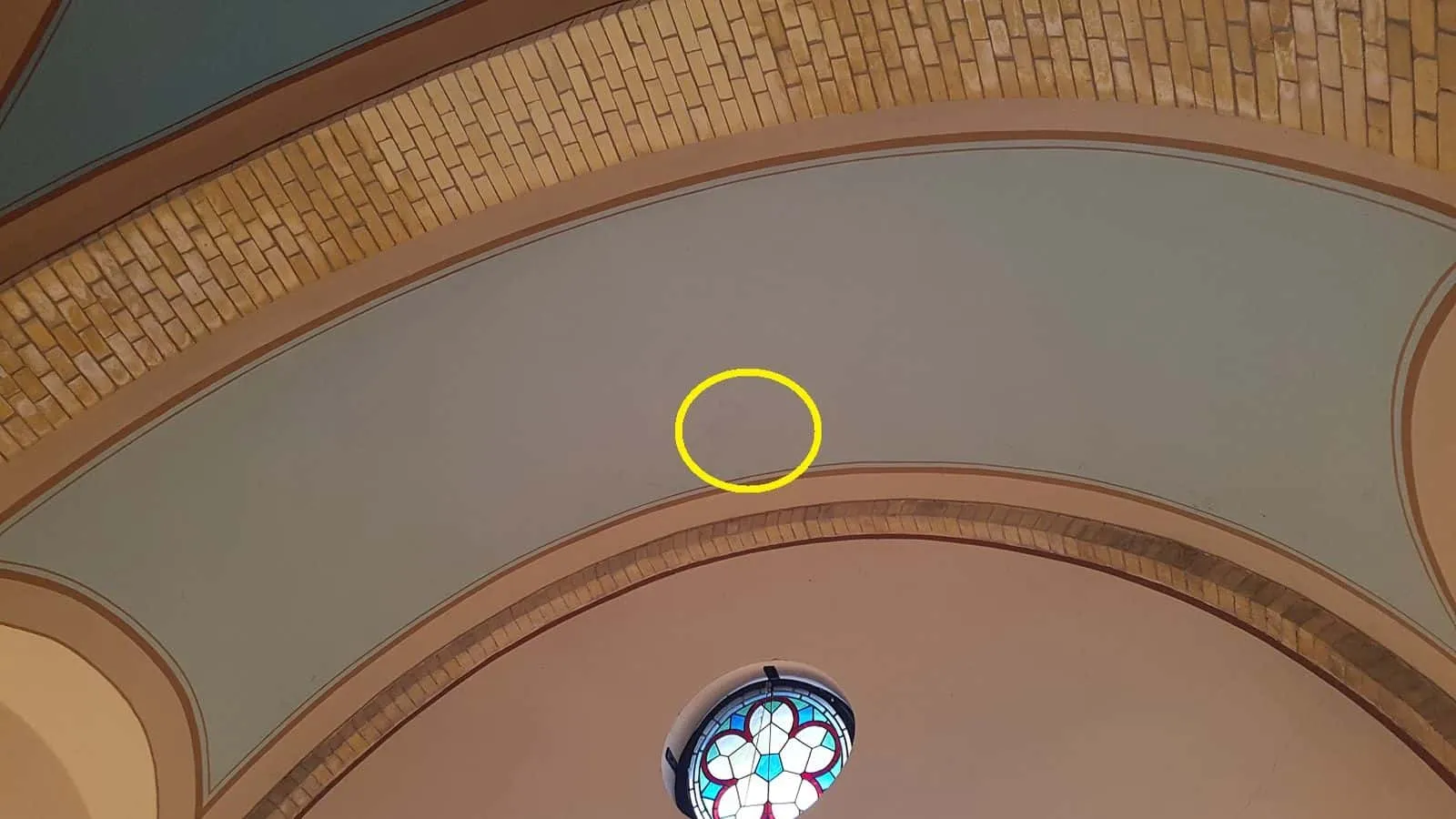 Gewölbte Decke mit gemauerten Bögen und Rundfenster, ein gelber Kreis markiert den Wasserfleck im Putz