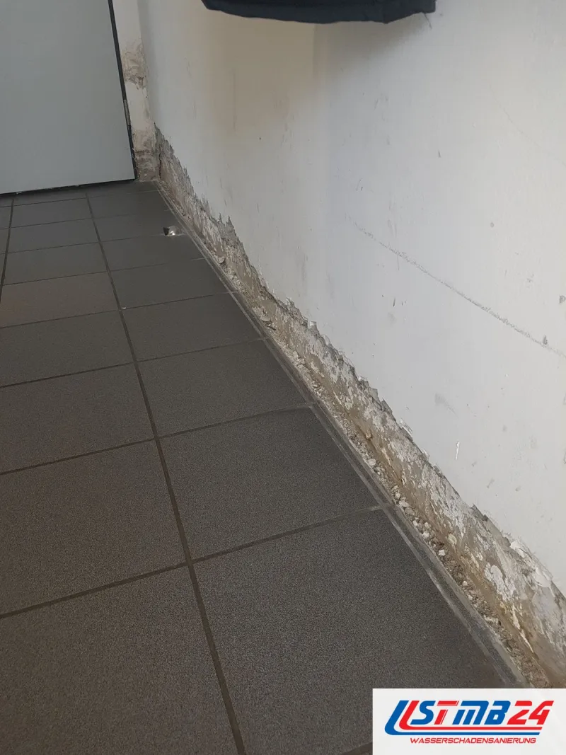 Wand über grauen Bodenfliesen, der Putz ist am Sockel streifenweise abgeschlagen, darüber zeichnet sich der feuchte Rand ab
