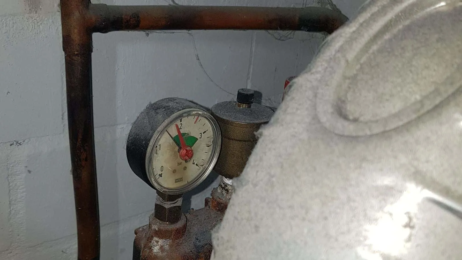 Manometer an einem alten Kessel, darüber rostige Rohre vor einer hellen Kellerwand