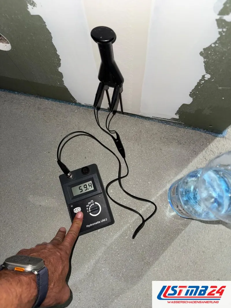 Feuchtemessgerät mit 59,9 auf dem Display auf dem Estrich, die Messkugel steht an der Wand mit abgeschlagenem Putz