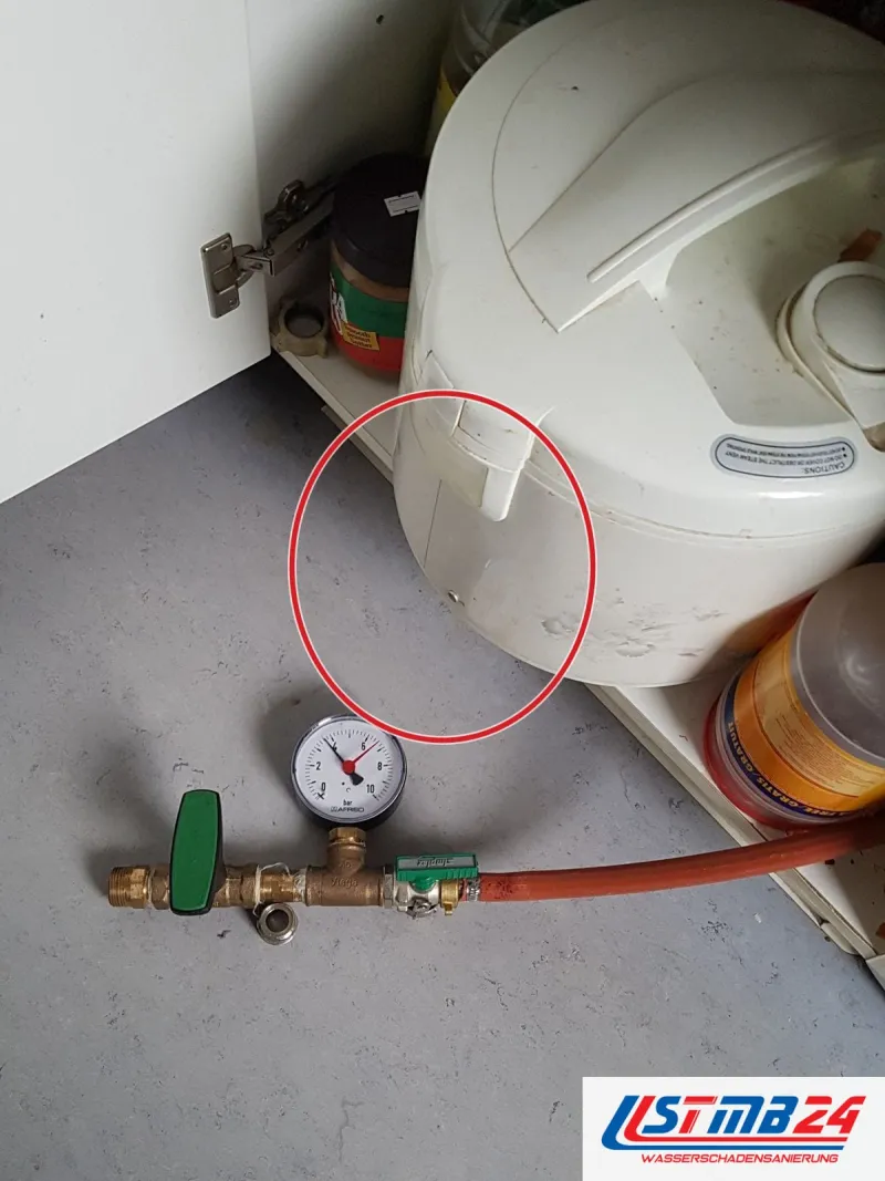 Prüfarmatur mit grünem Hahn und Manometer auf grauem Boden vor einem Unterschrank mit Reiskocher und Vorräten, ein roter Kreis markiert die Stelle