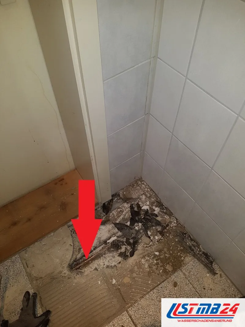 Aufgestemmter Boden in einer Badecke, ein roter Pfeil zeigt auf die freigelegte Kupferleitung zwischen Fliesenresten und Schutt