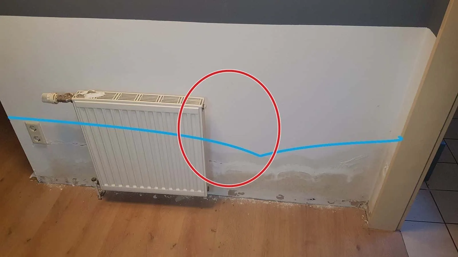 Heizkörperwand mit blauer Linie über Wasserrändern und Ausblühungen, die Sockelleiste ist abgenommen