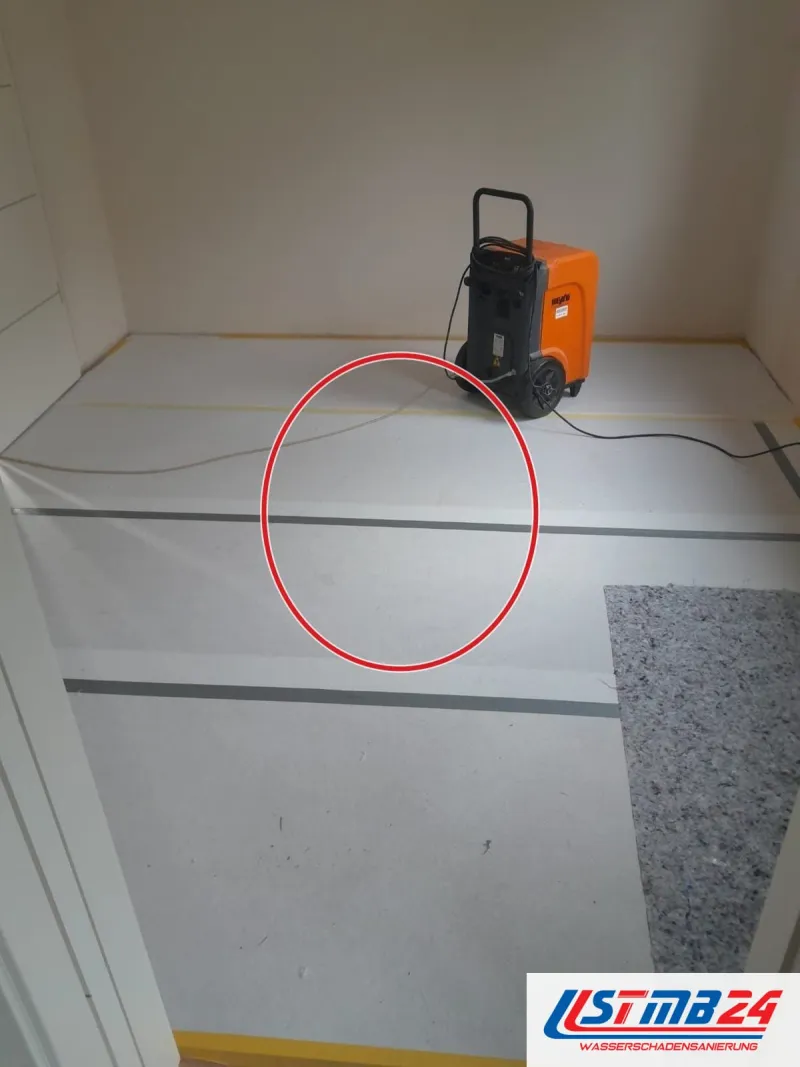 Oranger Kondenstrockner auf Abdeckpappe in einem leeren Zimmer, Vlies und Klebeband schützen den Boden, ein roter Kreis markiert die Stelle