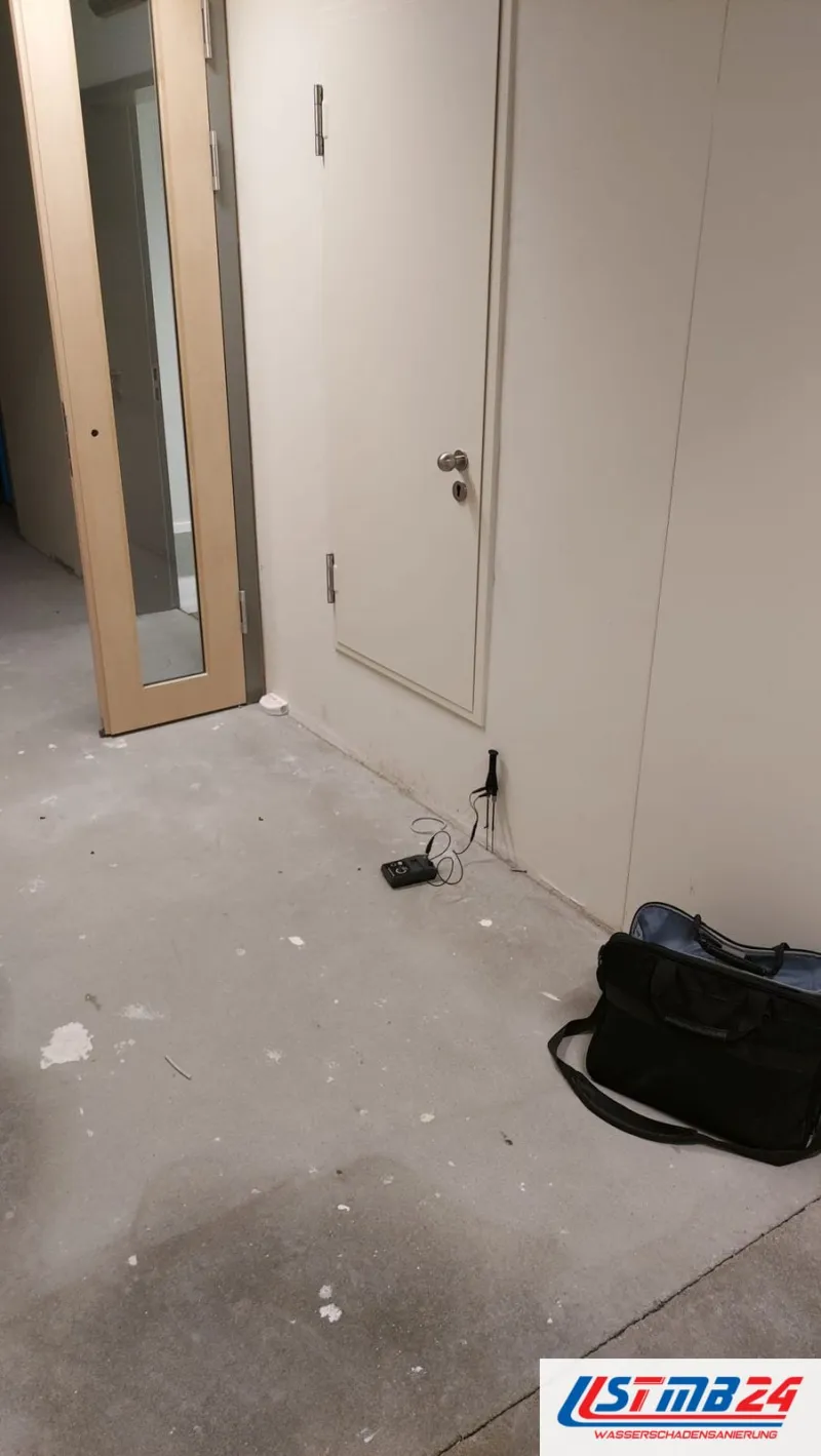 Leerer Raum mit freiliegendem Estrich, am Boden ein Messgerät mit Kabel und eine Gerätetasche, an der Wand lehnt eine Tür