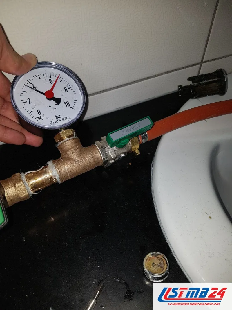 Manometer an einem Messingventil mit grünem Griff, in der Hand gehalten, daneben ein orangefarbener Schlauch und ein Waschbecken