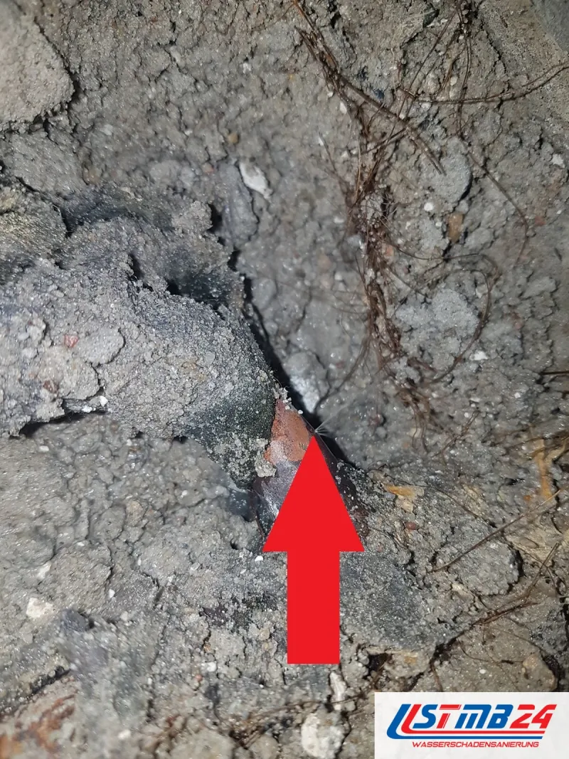 Aufgebrochener Boden mit Schotter und Erdreich, ein roter Pfeil zeigt auf die nasse Stelle an der freigelegten Leitung
