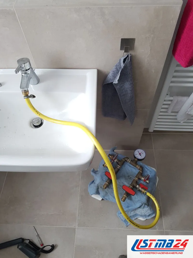 Waschtisch mit gelbem Prüfschlauch an der Armatur, auf einem blauen Tuch am Boden steht die Prüfarmatur mit Manometer und zwei roten Hähnen