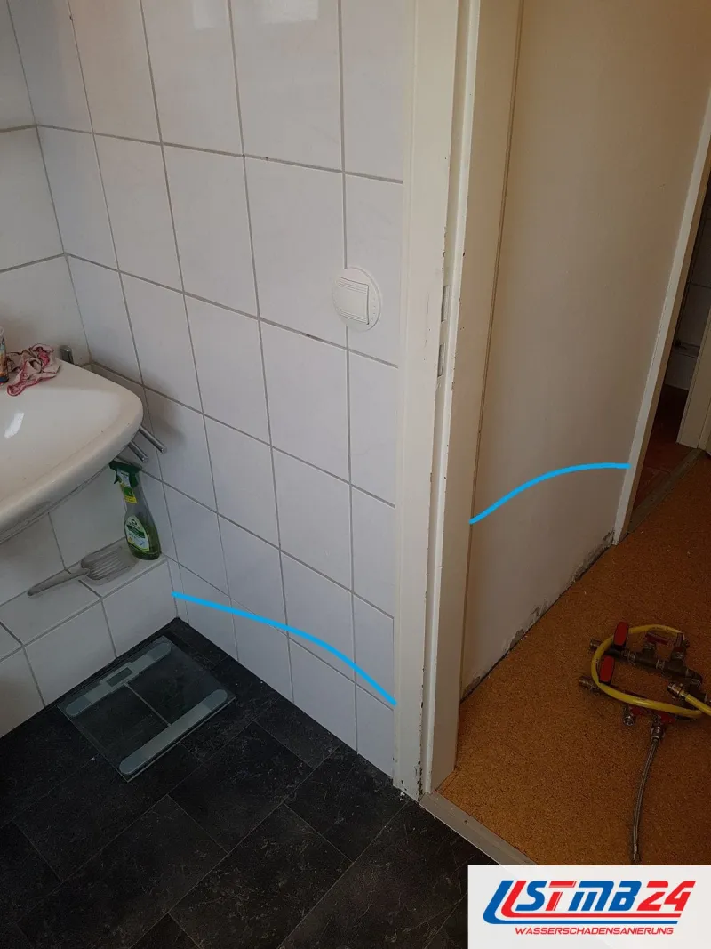 Blaue Linien zeichnen den Leitungsweg unter der Schwelle einer Badtür nach, links Wandfliesen, rechts Korkboden, im Flur die Prüfarmatur