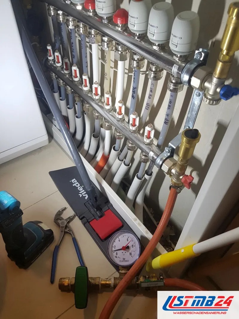 Heizkreisverteiler mit weißen Stellköpfen, davor ein Manometer am roten Prüfschlauch, Zange und Akkuschrauber auf dem Boden