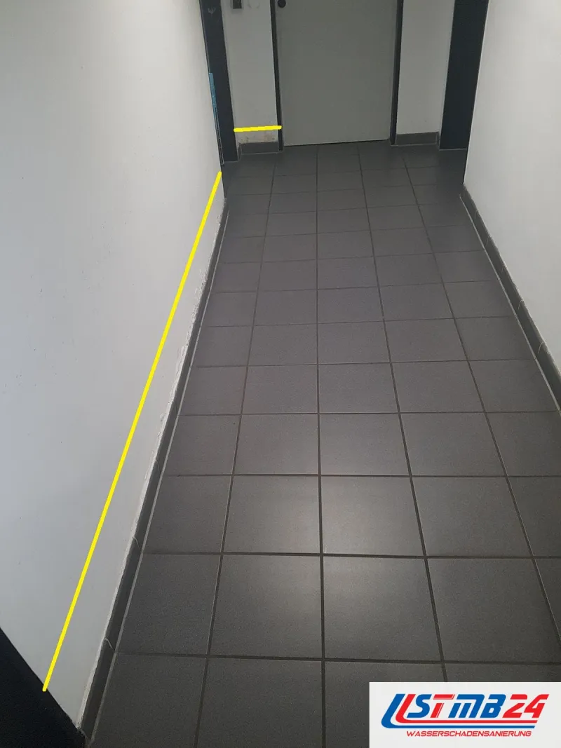 Langer Flur mit grauen Bodenfliesen, eine gelbe Linie am Wandfuß zeigt, wie weit die Feuchte an der Wand entlangläuft